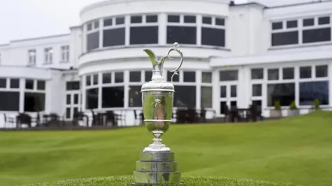 PA Media The Claret Jug is pictured on display on grass in front of the Royal Birkdale clubhouse, which is a large two-storey white fronted art deco building. Tables can be seen on a terraced area in front of the clubhouse.