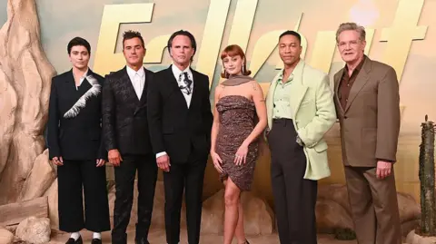 Getty Images Xelia Mendes-Jones, Justin Theroux, Walton Goggins, Ella Purnell, Aaron Moten e Kyle MacLachlan estão em frente a um quadro que diz Fallout em letras douradas. 
