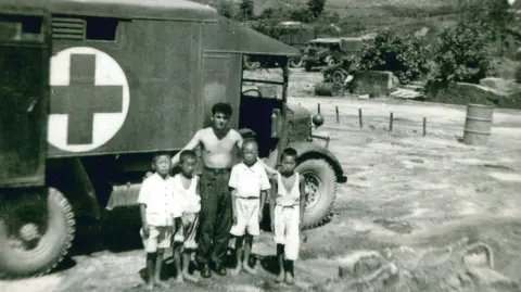 Albert Morrow Albert Morrow centre, with four children wearing light coloured clothing, in front of an ambulance van