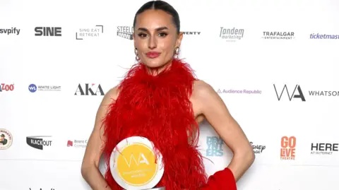 Getty Images Amber Davies, holding her circular yellow WhatsOnStage award. She has a brunette slick-back bun, wearing a red dress and big pearl earrings. She stands in front of a branded white wall with sponsor logos.