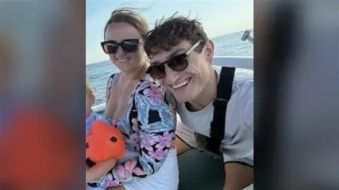 Demie Risby A blonde woman and a man with dark curly hair taking a selfie. They both have dark sunglasses on and are sitting on a boat.