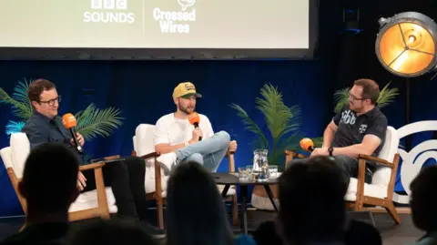 Three men sit on white chairs in front of an audience. They are holding black microphones with orange covers. 