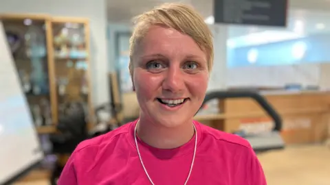 A woman with short blonde hair and wearing a pink shirt, smiles as she stands inside a building.