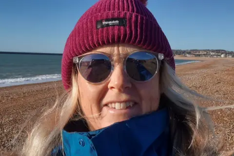 Julie A close up of a woman with light blonde hair at the beach. She wears a maroon beanie and a blue raincoat with a high collar, as well as sunglasses. 