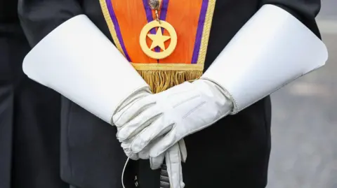 Getty Images A person in white gloves holding the top of a sword. The person is wearing an orange sash and dark suit