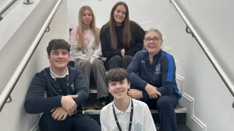 BBC The photo shows a group of five people sitting together on an indoor staircase. Three individuals are seated on the steps toward the back, and two are positioned lower down. The staircase has smooth white walls on both sides with metal handrails running along the length of the stairs. Some of the group are wearing clothing with school emblems, including dark jumpers, sports-style tops, and a white polo shirt