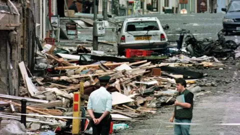 PA Media Archive photo of the aftermath of the Omagh bomb in August 1998. Piles of wood and rubble lie on the street outside damaged shops. The wreckage of a car and a damaged wheelchair are in the background. A uniformed RUC office stands at a cord with his hands behind his back. Another man wearing a headset and holding a book is talking to the officer.