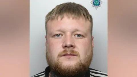 Staffordshire Police A mugshot of Tommy McQuaker. He is wearing a black jacket with white stripes on the shoulders. He has short blonde hair and light brown beard and moustache