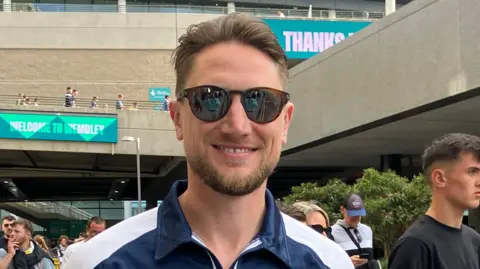 Henry Godfrey-Evans/BBC David Bonnett wearing sunglasses and a navy Southend top. The stadium and lots of fans are behind him.