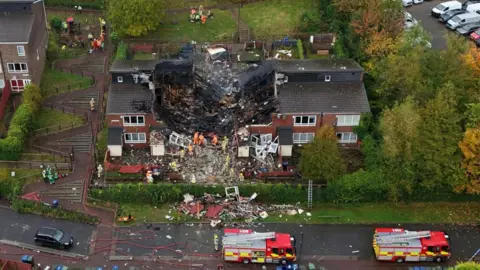 The house following the explosion. A large crater shaped hole is missing from the rood. Debris from the building is scattered on the nearby grass and  street. Two fire engines are parked on the road and fire fighters are clearing rubble from the building.