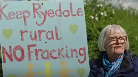 A woman with grey hair and glasses holds up a placard reading 'keep Ryedale rural - no fracking.' 