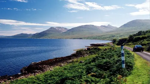 A car travels down a road, by the sea, on the Isle of Mull. The car is heading towards some mountains. 