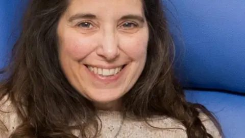Simone Hunter has long dark hair and is sitting in a blue hospital chair.
