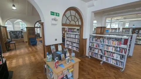 North Northamptonshire Council An inside of a library, with a wooden arch, books on lots of shelves, a wooden herringbone floor, tables, and arched ceilings. 