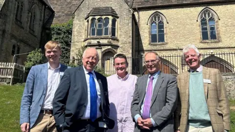 A mixed group of formally dressed men stand in front of a historic stone building.