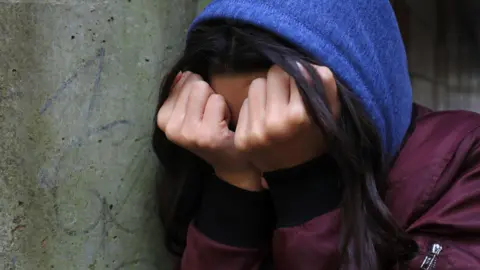 PA Media A girl with dark hair holds her hands over her face to cover it from the camera. She is wearing a maroon dark red jacket over a blue hooded jumper, with the hood pulled over her head. She is leaning against a concrete wall which has graffiti on it.