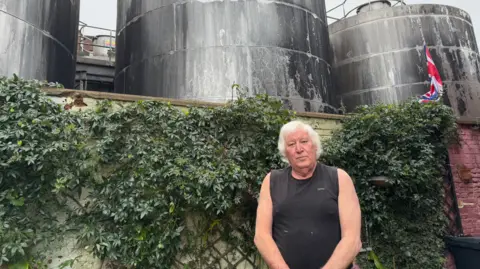 George King/BBC An elderly man wearing a black vest standing in front of a garden wall and a brewery