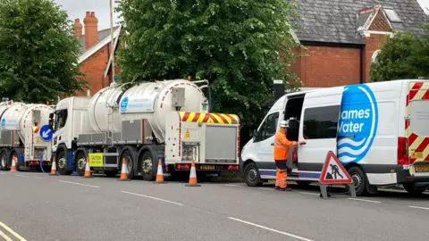 A white branded Thames Water van parked behind two large water tanker trucks. There are orange cones and road signs advising drivers of the disruption. A utility worker wearing orange hi-vis is closing the sliding door of the van. In the background there are trees and red brick houses with slate roofs. 