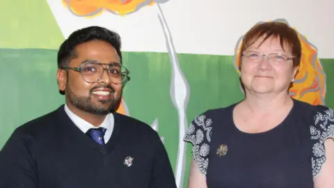 Christy Vijay and Helen Simpson are smiling at the camera. Christy is wearing large glasses and has a navy tie on under a sweater. Helen is wearing wire-rimmed glasses and a top with frilly sleeves.