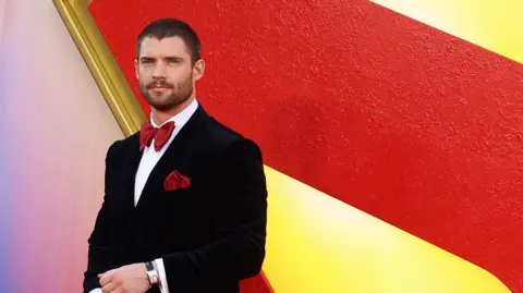 David Corenswet in a tux and red bowtie in front of a huge S in the red and yellow colours
