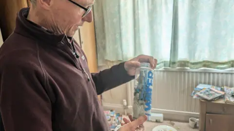 Andrew holds up a water bottle crammed with syringes. In the background is his son's bedroom, with dirty plates and litter strewn around.