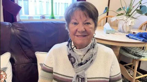 A woman sitting on a sofa with short dark hair looking into the camera. She has a cream jumper and is wearing a grey scarf. In the background is a table and chairs.