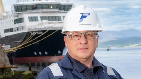 FMPG A bespectacled man in blue overalls and a white hard hat. The large ferry Glen Rosa can be seen in the background