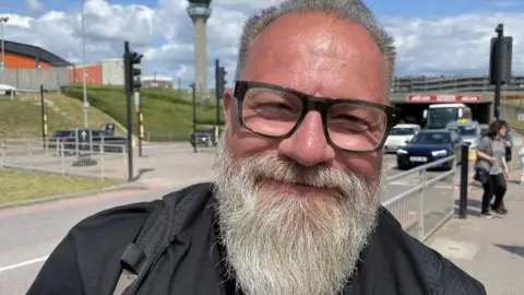 Helen Mulroy/BBC Chris Smith has swept-back hair and a beard. He is wearing glasses, and a black top and rucksack. He is standing outside with a road and air traffic control tower in the background.