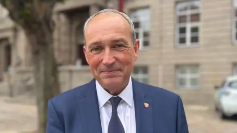 Senator Lyndon Farnham, stands in Royal Square wearing a navy suit, white shirt and dark tie. He is positioned in the foreground, with a large tree and historic stone-fronted buildings softly blurred in the background. The square’s paved surface, a parked vehicle and a few distant figures are visible around the edges of the scene.