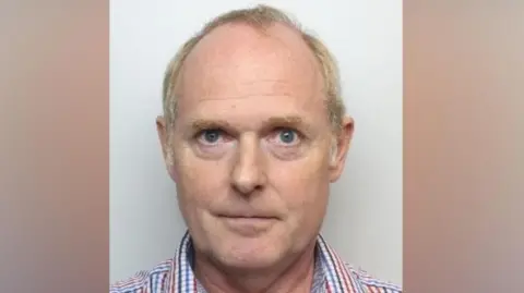 Mugshot of Christopher Cave. He has short blonde grey hair. He is wearing an open-necked, checked shirt.