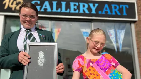 Jon Wright/BBC Two young people stand in front of a shop window. The sign at the top reads "142 gallery.art". They are both holding up pictures, one is a white chalk design on black paper and the other is brightly coloured squares in pink and orange