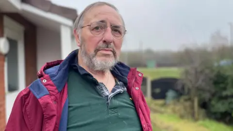 Simon Thake An elderly man with glasses dressed in a purple jacket and green shirt stands outside looking impatient