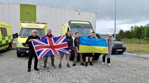 Oliver Ansell-Hodges Six people - five men and one woman - are standing in front of parked yellow ambulances. Between them they are holding a Union Jack flag and a Ukrainian flag.