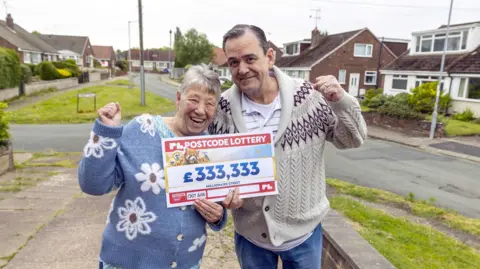 A couple hold up a cheque for £333,333 with their fists in the air. Nancy is wearing a light blue cardigan with white flowers across it. Colin is wearing a cream coloured cardigan with a maroon pattern on the upper section.