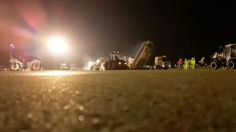 Resurfacing equipment on the tarmac of an airport runway. The shot is taken from a low angle so you can see the tarmac, the machines, workers in yellow high vis and the sky at night. It's all lit up by a big light in the air. 