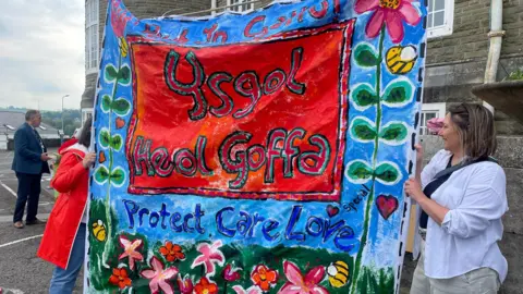 protestors hold up a mural sign says Ysgol Heol Goffa protect care and love