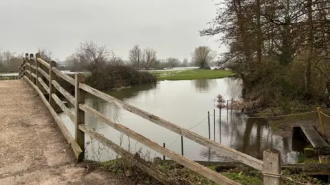 Luke Deal/BBC A concrete pedestrian bridge with wooden fencing, with a flooded waterway underneath. The fields in the distance are also partially flooded.