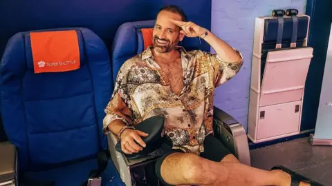 Jay Robert Jay Robert, a man wearing a patterned shirt, shorts and sandals, sit on a plane seat and salutes while smiling