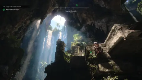 Ubisoft A screenshot. The main character is seen from a distance climbing rocks inside a cave. There is an opening near the top from which water is pouring, with light coming through too. It creates an effect of the scene being cinematically lit - a trick modern games often utilise.