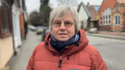 Woman wearing circular glasses, red puffer coat and blue scarf posing for a photo in front of a blurred suburban street with a school and houses in the background