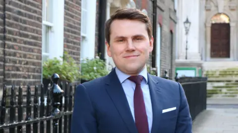 Taxpayers' Alliance A person in a navy suit and burgundy tie stands on a historic street lined with Georgian brick buildings and black railings.