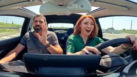 Two people in a car; the woman driving looks surprised