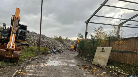 LDRS A digger is parked up at the entrance to the rubbish dump. Large mounds of rubbish and tyres can be seen.