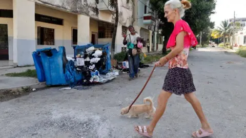 EPA/Shutterstock A woman dressed in dark shorts and a light floral top walks with a dog on a lead past a large blue rubbish bin in Havana burned the night before in protest against prolonged blackouts. A man in jeans and T-shirt is seen in the background. 
