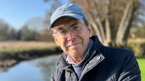 Tom Jackson/BBC Colin Day is next to a small river. He is wearing a light blue baseball cap, a tartan shirt and a navy blue jumper and coat. 