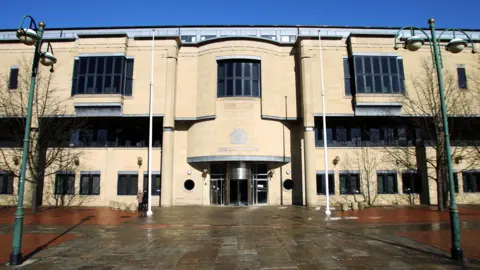 The front of Bradford Crown Court, a multi-storey building made of sand-coloured brick. The words "The Law Courts" as well as a crest can be seen above the main entrance.