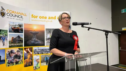 Lee Trewhela / LDRS Kate Ewert is pictured giving a speech at the Bodmin Leisure Centre election count on Friday, 2 May 2025. She is wearing a black dress and has a red ribbon and is stood in front of a Cornwall Council sign. 