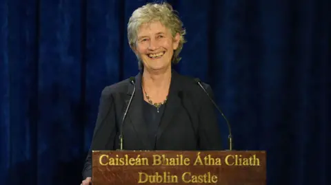 Niall Carson/PA Media Catherine Connolly stands behind a dark wooden podium and smiles. There are two microphones placed in front of her. She is standing in front of a long, deep blue velvet curtain. In gold inscription, the podium says 'Caisleán Bhaile Átha Cliath Dublin Castle' on it. Connolly is wearing a dark grey suit jacket and a dark shirt underneath.