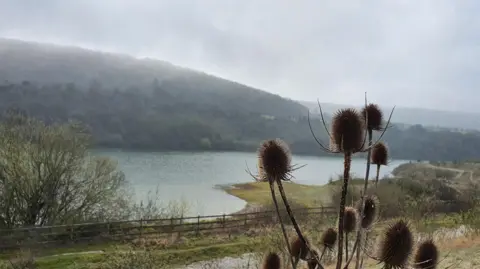 boxerbabe A lake backed by a hill. A fenced off path is in the foreground.