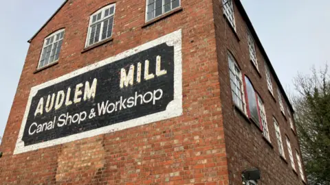 The side view of a tall red brick building, with the words Audlem Mill Canal Shop and Workshop painted in black and white below a row of windows